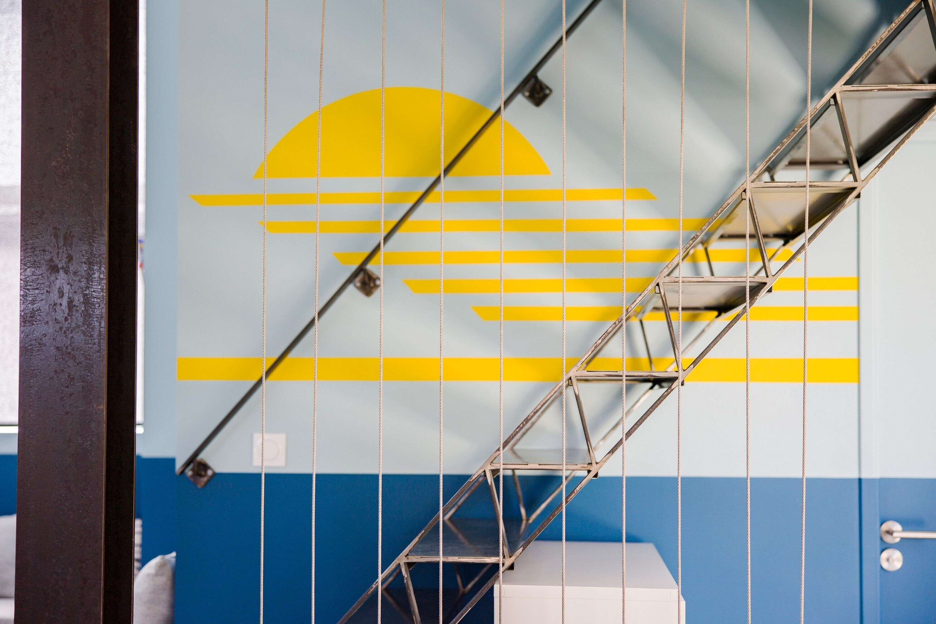 A metal staircase with a geometric design, a blue and yellow wall with a sun motif, and a white object at the bottom.