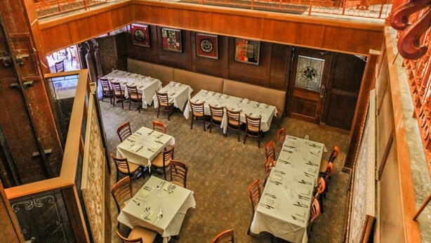 A restaurant with a high ceiling featuring stained glass, a second-floor balcony with ornate railings, and multiple dining tables set for guests.