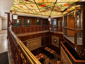 Eine prachtvolle Halle mit hoher Decke, die farbenfrohe Glasfenster, eine Holztreppe mit aufwendiger Geländerarbeiten und einen Essbereich mit gedeckten Tischen zeigt.