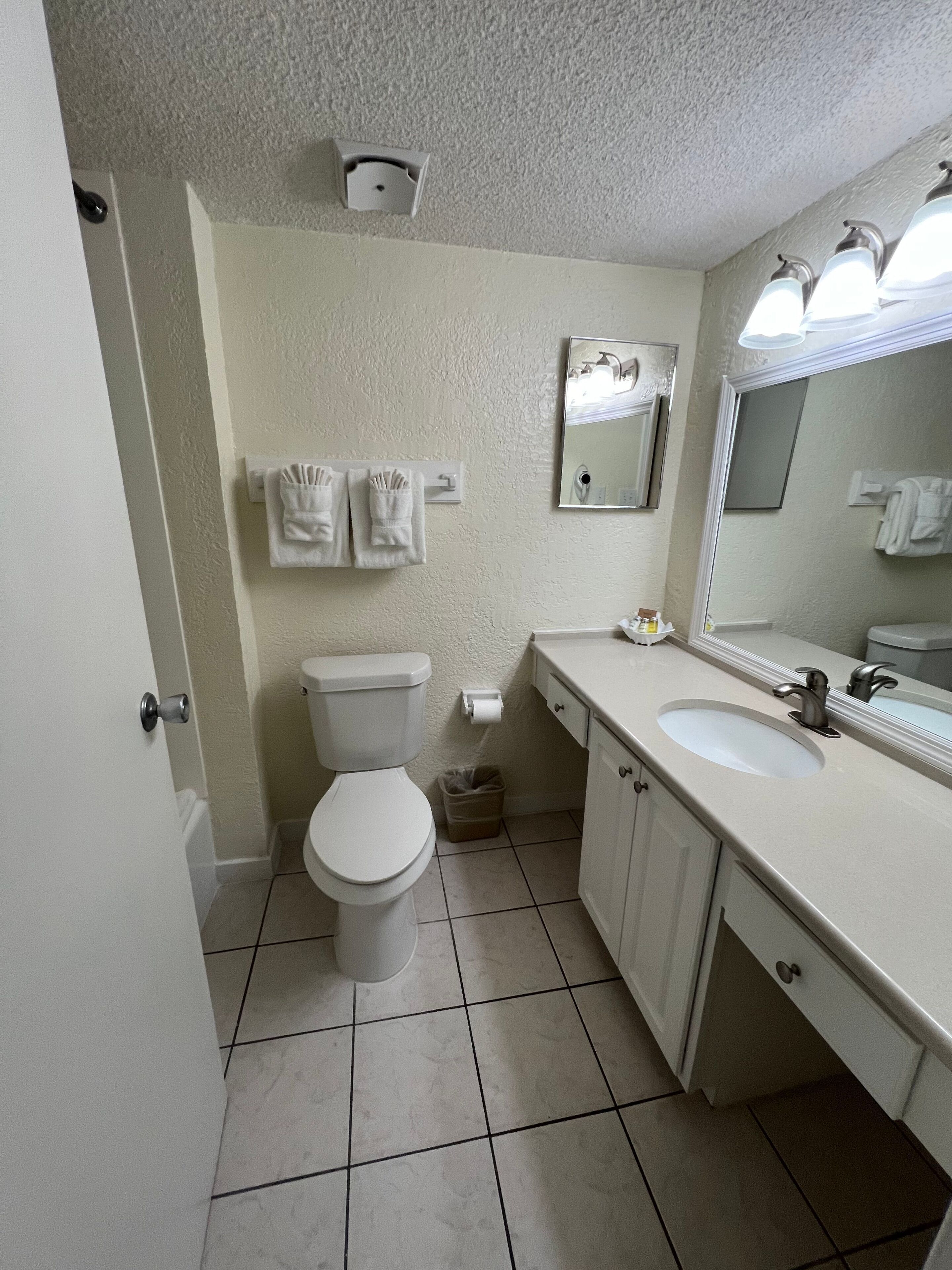 Une salle de bain avec des toilettes, un lavabo et un miroir. Il y a des serviettes accrochées au mur et une poubelle.