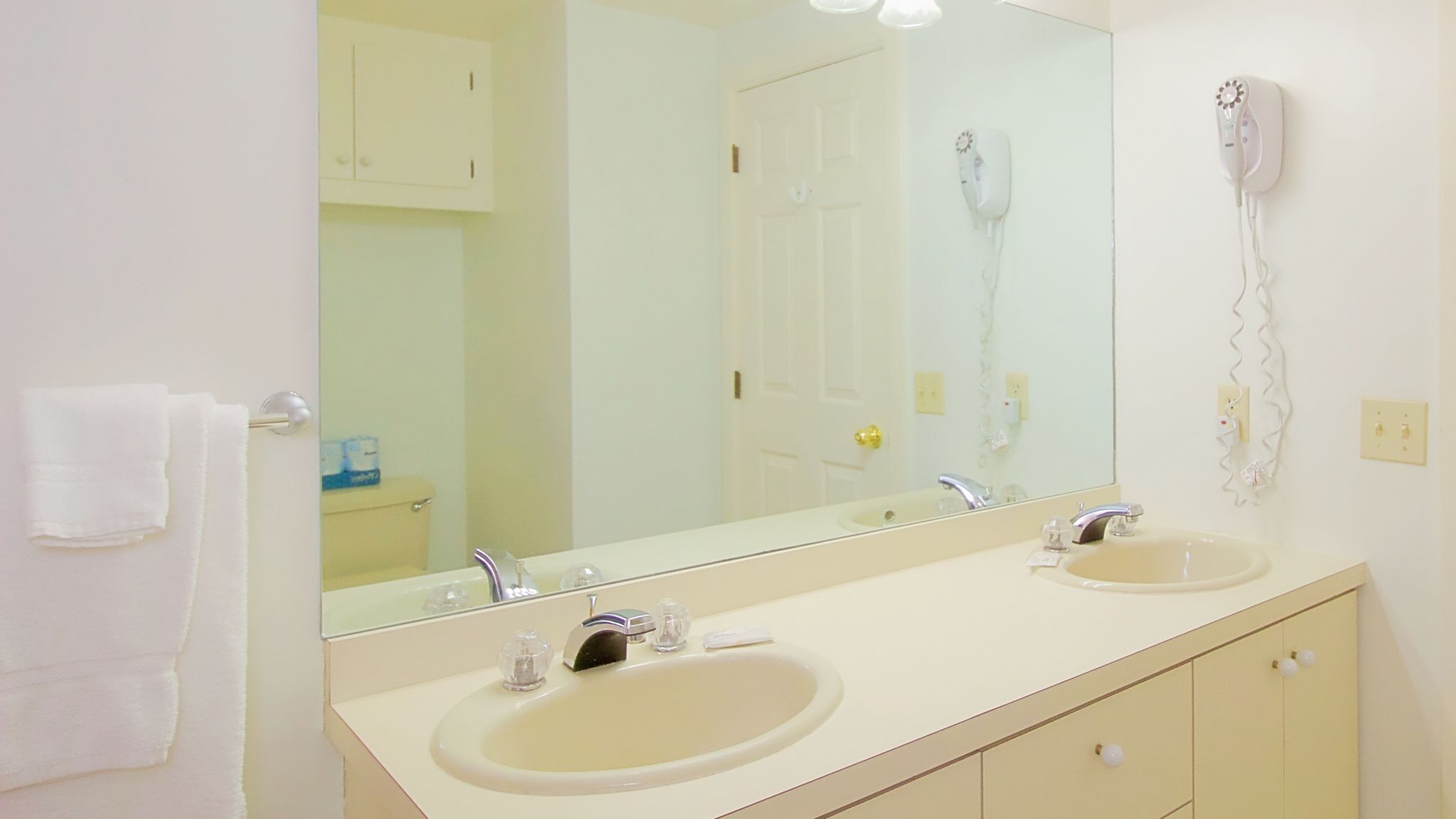 A bathroom with two sinks, a large mirror, a hairdryer on the wall, and a toilet visible through the door.