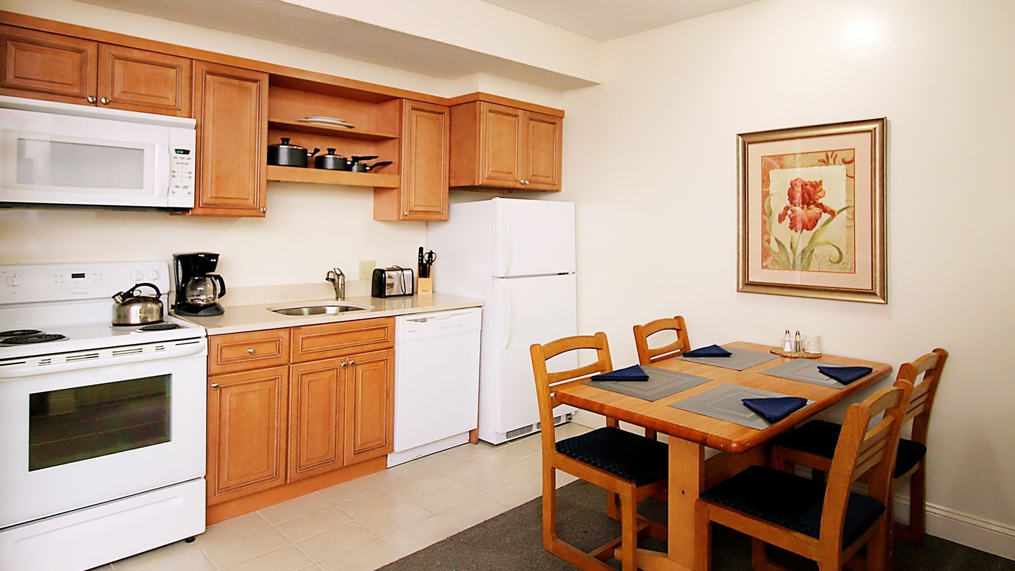 A compact kitchen with wooden cabinets, a microwave, stove, and a refrigerator. There is a dining table with chairs and a framed picture on the wall.