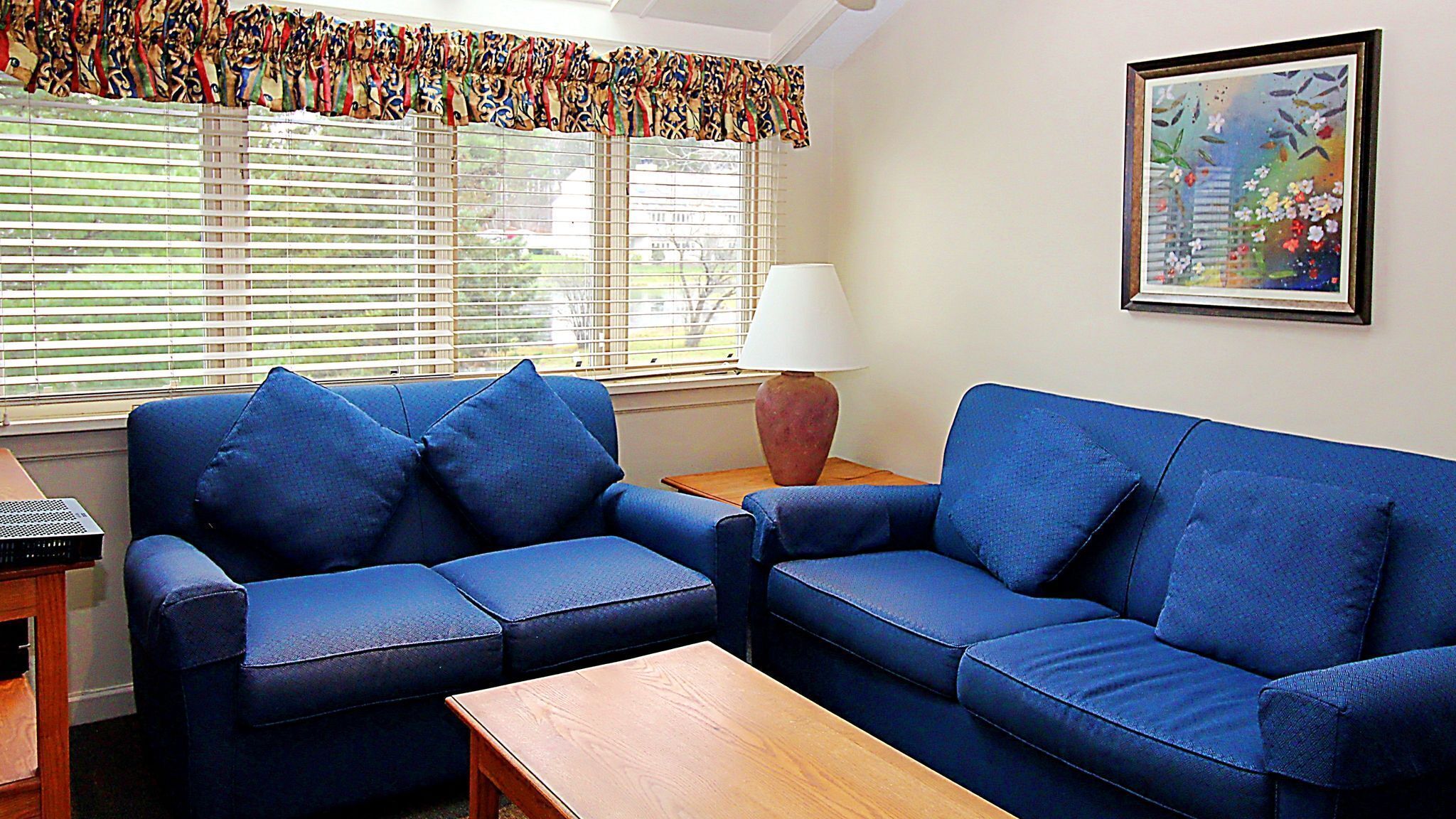 A living room with a blue sofa, a wooden coffee table, a lamp, and a window with blinds.