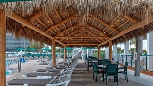A covered outdoor seating area with lounge chairs and tables, a pool in the background, and a view of a building across the pool.