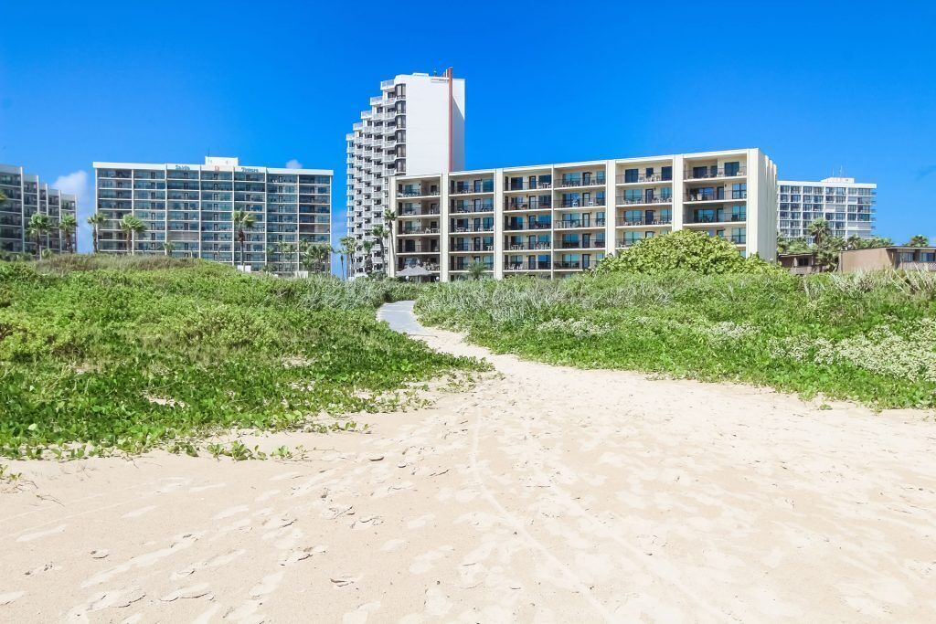 A beachfront hotel with multiple buildings, a sandy path leading to the beach, and lush greenery.