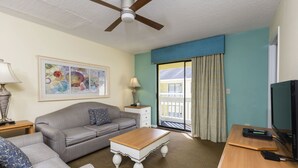 Habitación de hotel con ventilador de techo, un sofá, una mesa de centro, un televisor y una ventana con cortinas.