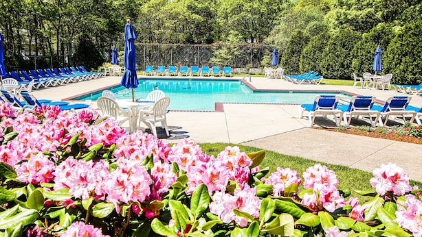 A pool area with blue lounge chairs and white chairs, surrounded by pink flowers and trees.
