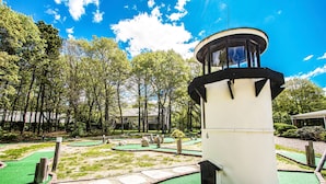 A lighthouse structure with a circular observation deck, surrounded by a green artificial turf area with stepping stones and a small pond.