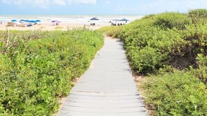 A paved pathway leading to a beach with umbrellas and people in the distance.