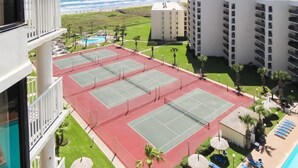 A tennis court surrounded by a pool, palm trees, and a beach view.