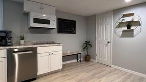 A modern kitchen with white cabinets, a stainless steel dishwasher, and a microwave. There is a bench and a potted plant in the corner.