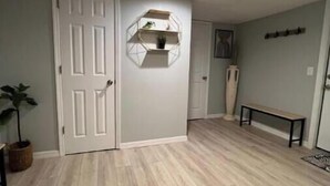 A hallway with wooden flooring, a white door, and a hexagonal shelf with a plant.