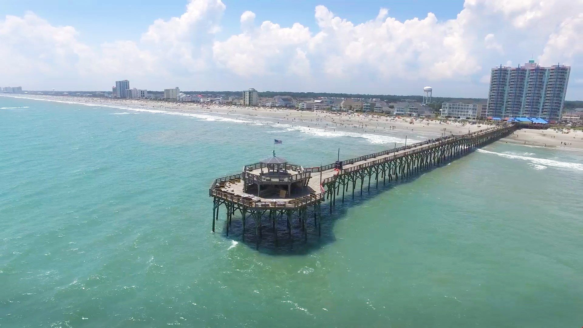 Un muelle se adentra en el océano, con un pabellón al final, rodeado por una playa y edificios al fondo.