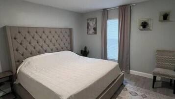 Un dormitorio con una cama grande, un cabecero, una ventana con cortinas, una mesita y una silla.