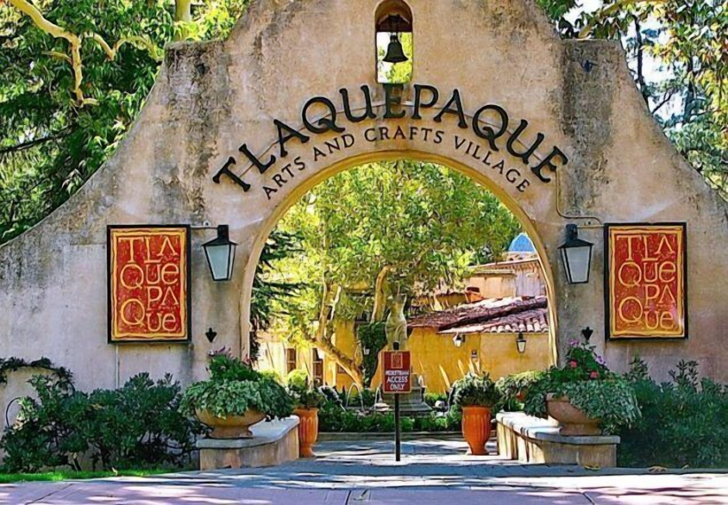 The entrance to La Que Paque Arts and Crafts Village with a stone archway, potted plants, and a sign indicating visitor access.