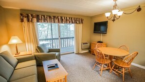 A cozy living room with a sofa, a coffee table, and a dining table with chairs. There is a TV mounted on the wall and a chandelier hanging from the ceiling. - 1-Bedroom Condo near Lake Wallenpaupack (Hawley)