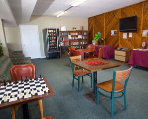 A room with a chess table, a vending machine, bookshelves, a television, and a table with a purple tablecloth.