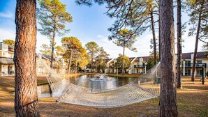 A hammock between trees overlooking a pond with buildings in the background.
