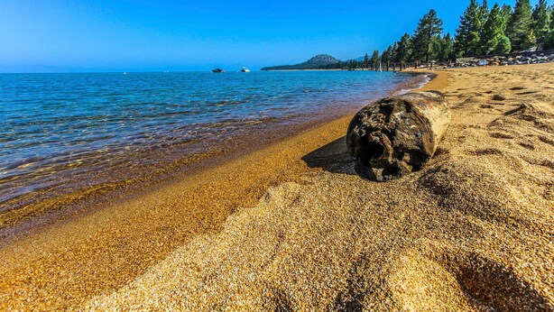 Una playa con agua cristalina, una roca grande en primer plano y una colina boscosa al fondo.