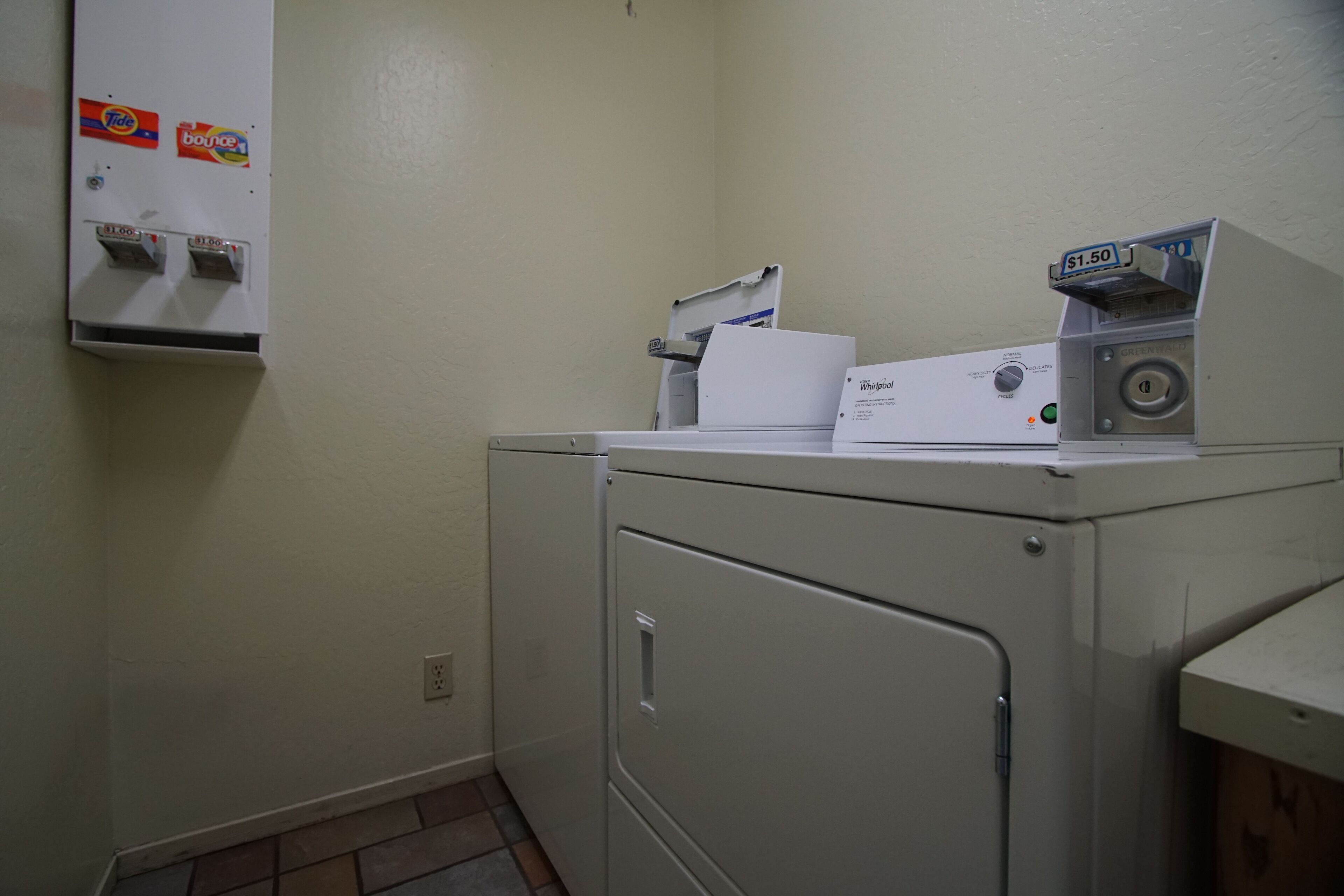 A laundry room with a Whirlpool washer and dryer, a coin-operated dryer, and a wall-mounted utility box with Tide and Bounce detergent dispensers.