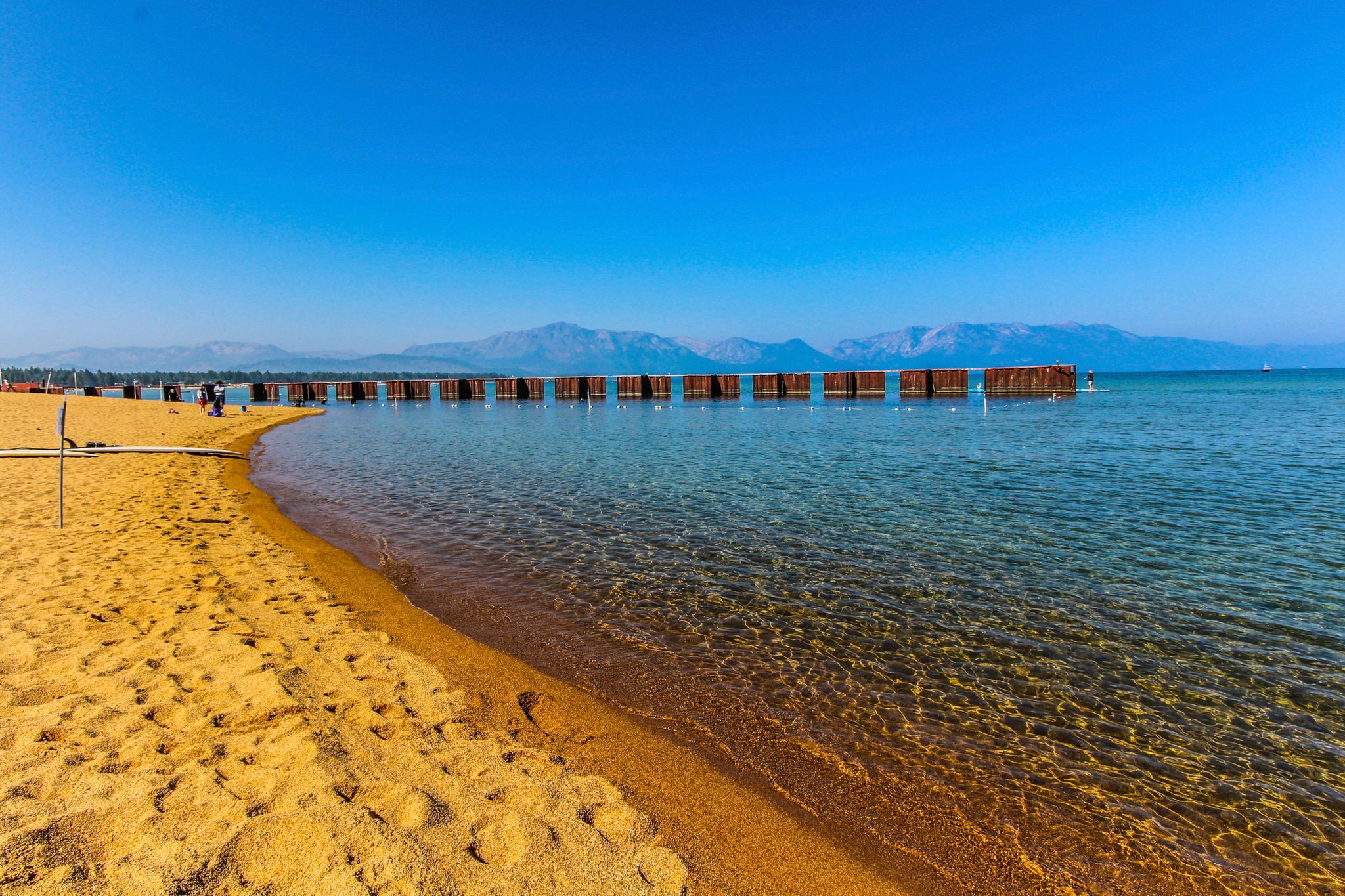 Una playa con agua cristalina, muelles de madera y montañas al fondo.