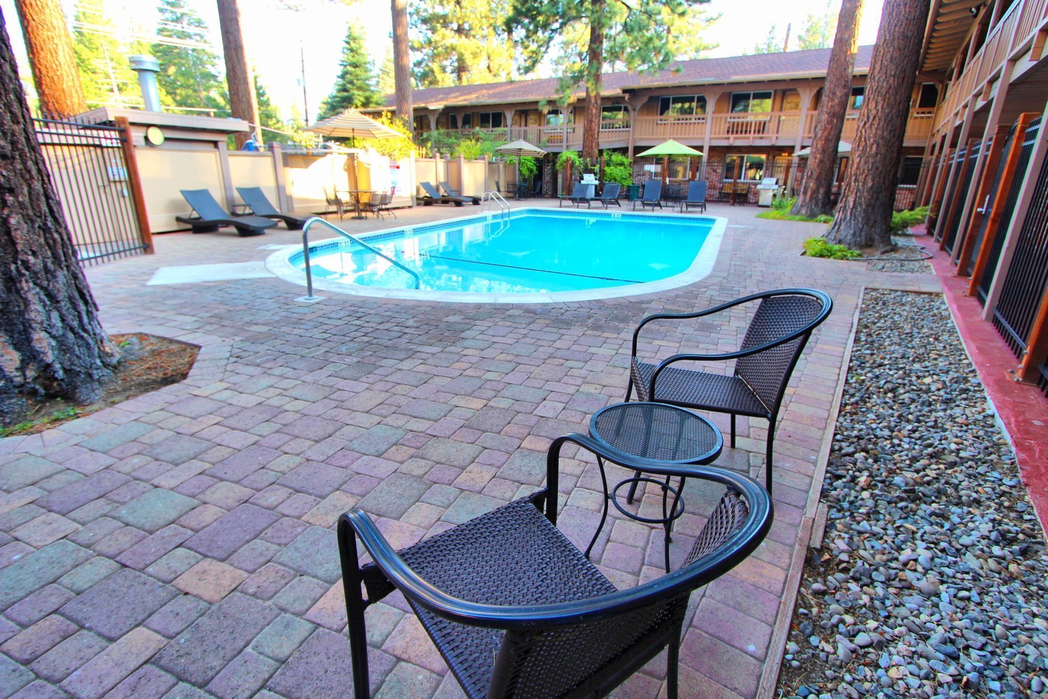 A pool area with lounge chairs and a table, surrounded by trees and a building with balconies.