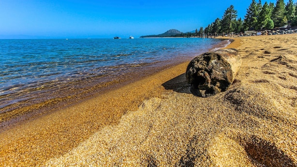 Una playa con agua cristalina, una roca grande en primer plano y una colina boscosa al fondo.