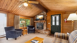 A cozy cabin interior with a fireplace, wooden walls, a ceiling fan, and a plaid sofa.