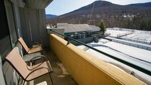 A balcony with two chairs, a view of a snow-covered area, and a building with a green roof in the background. - Condo Nr Loon Mountain | King Bed + Balcony Views (Lincoln)