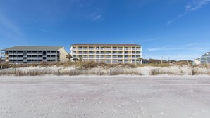 Ein mehrstöckiges Hotel mit flachem Dach und Balkonen, gelegen an einem sandigen Strand mit spärlicher Vegetation.