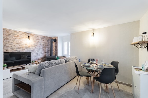 A modern living room with a flat-screen TV, grey sofa, dining table with chairs, and a brick accent wall.