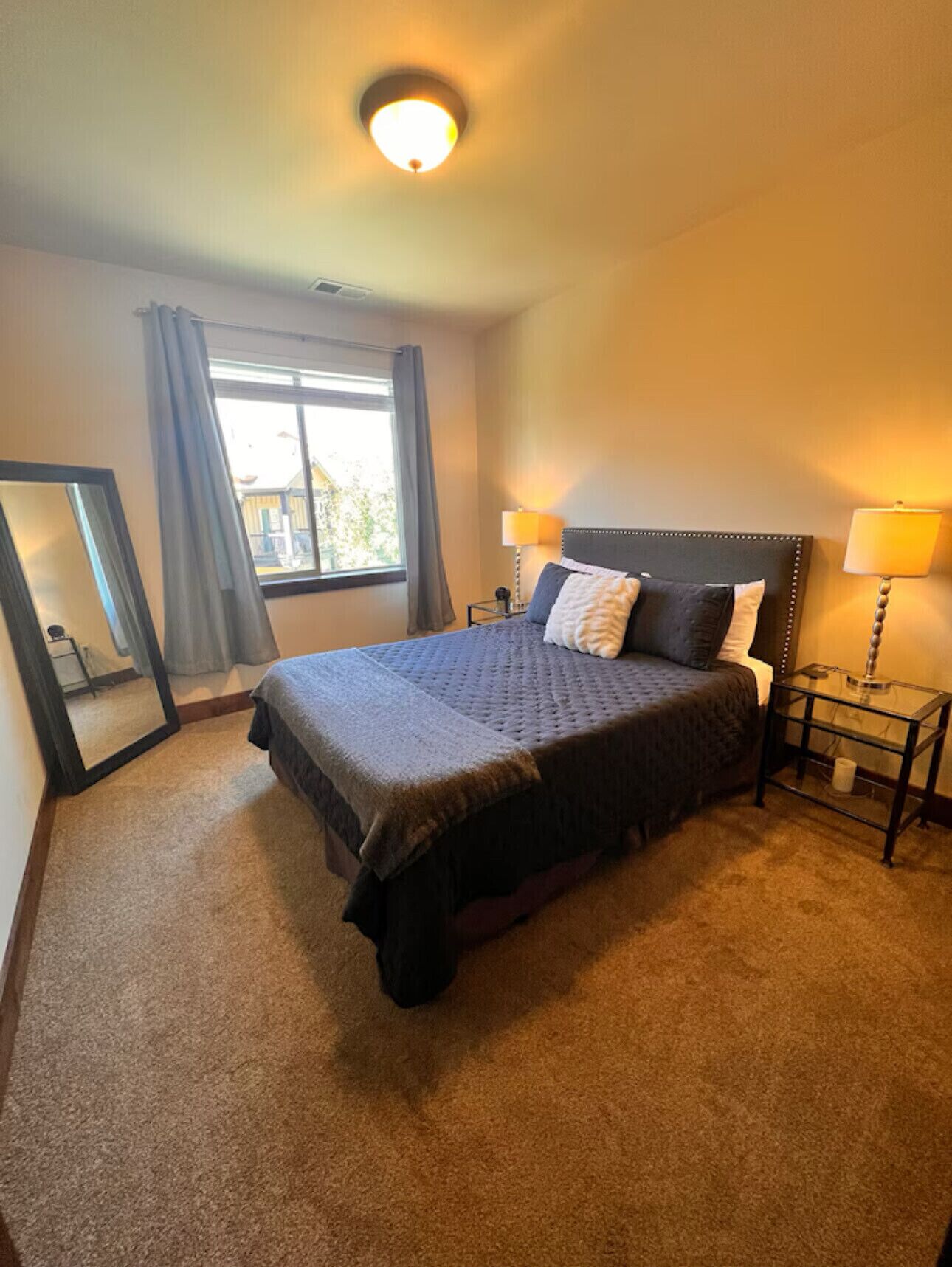 A neatly arranged bedroom with a large bed, two bedside lamps, a window with curtains, and a mirror on the wall.