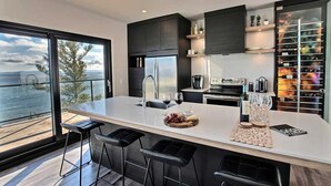 Una cocina moderna con una isla grande, taburetes de bar, vistas al océano y una estantería para vinos.
