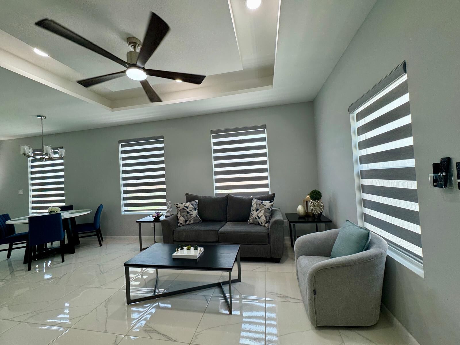 A modern living room with a ceiling fan, a dining area with a table and chairs, and large windows with blinds.