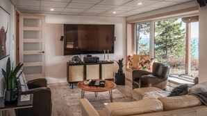 Een moderne woonkamer met een grote flatscreen televisie, een rieten dressoir, leren banken en uitzicht op de natuur.