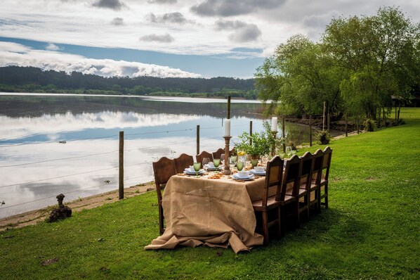 A lakeside dining setup with a table set for four, surrounded by chairs, and overlooking a calm lake with a forested backdrop.