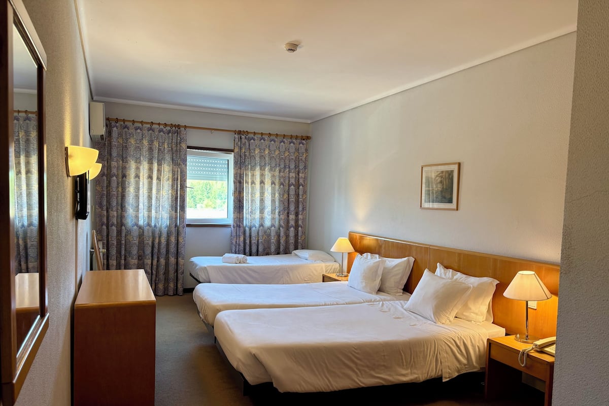 A hotel room with two beds, a desk, a chair, a window with curtains, a painting on the wall, and a ceiling light.
