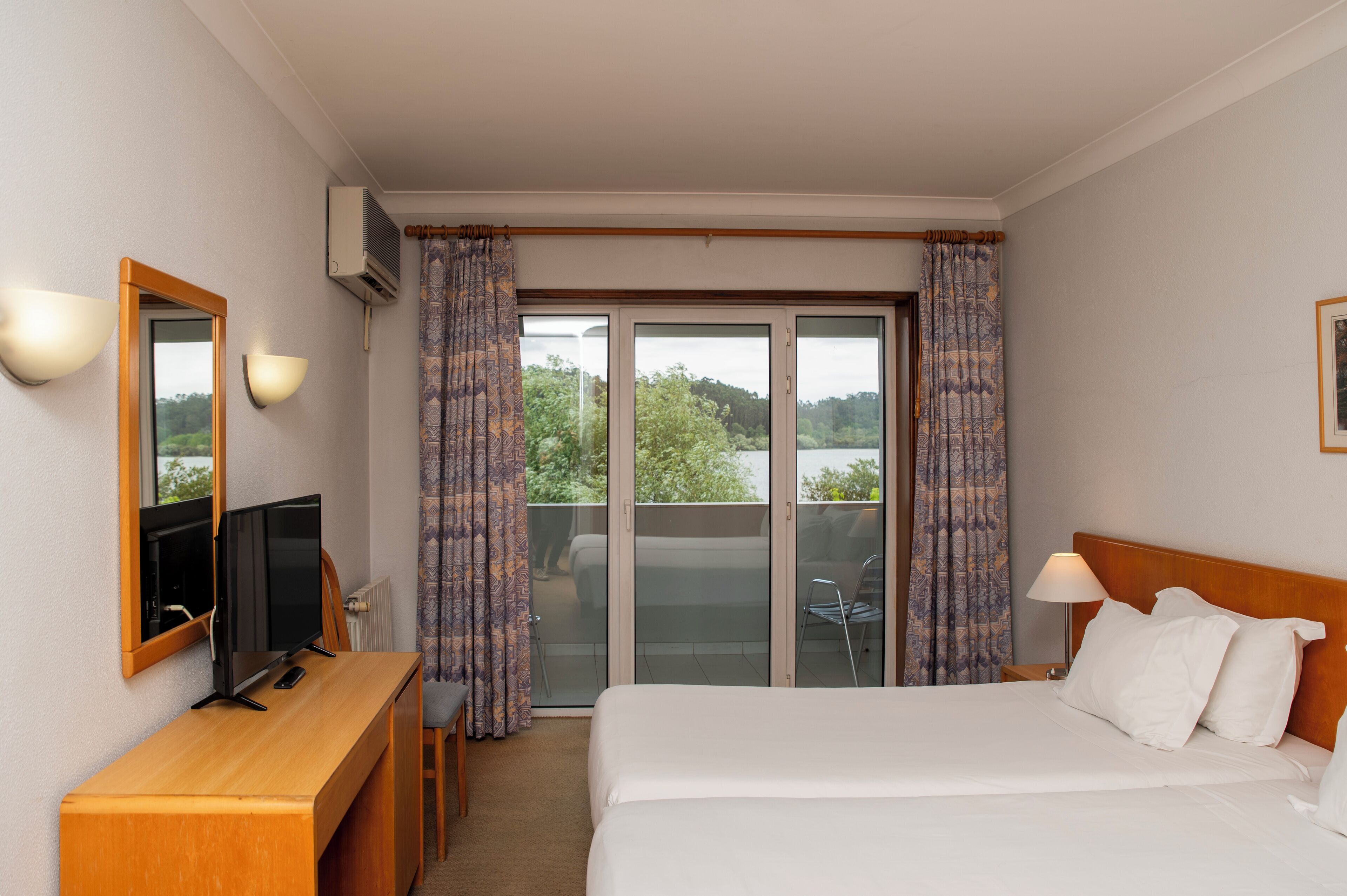 A hotel room with a bed, a desk with a computer, a TV, and a balcony with curtains.