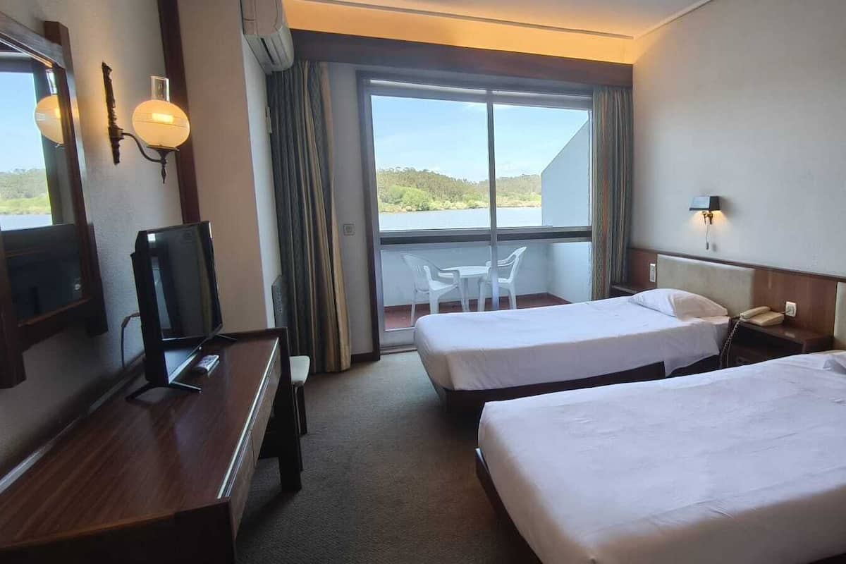 A hotel room with two beds, a desk with a TV, and a balcony with a view of the lake.