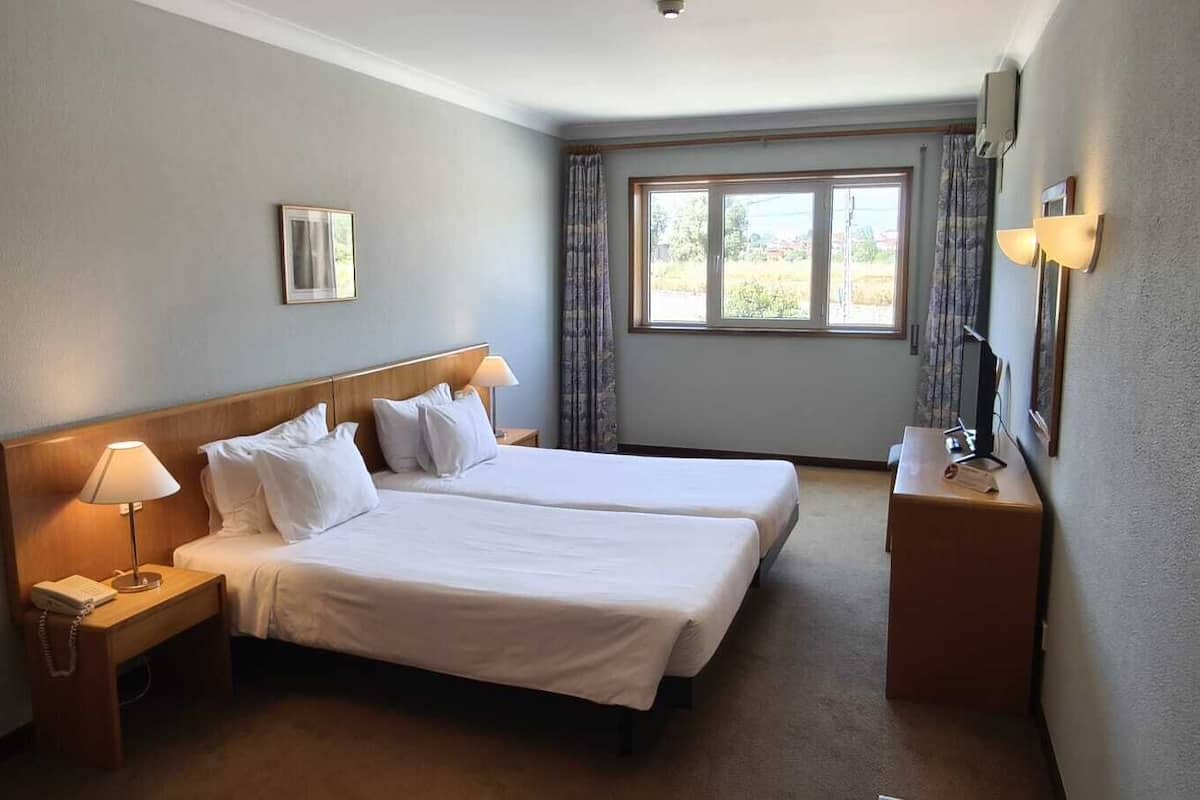 A hotel room with two beds, a nightstand with a lamp, a small table with a lamp, and a window with curtains.
