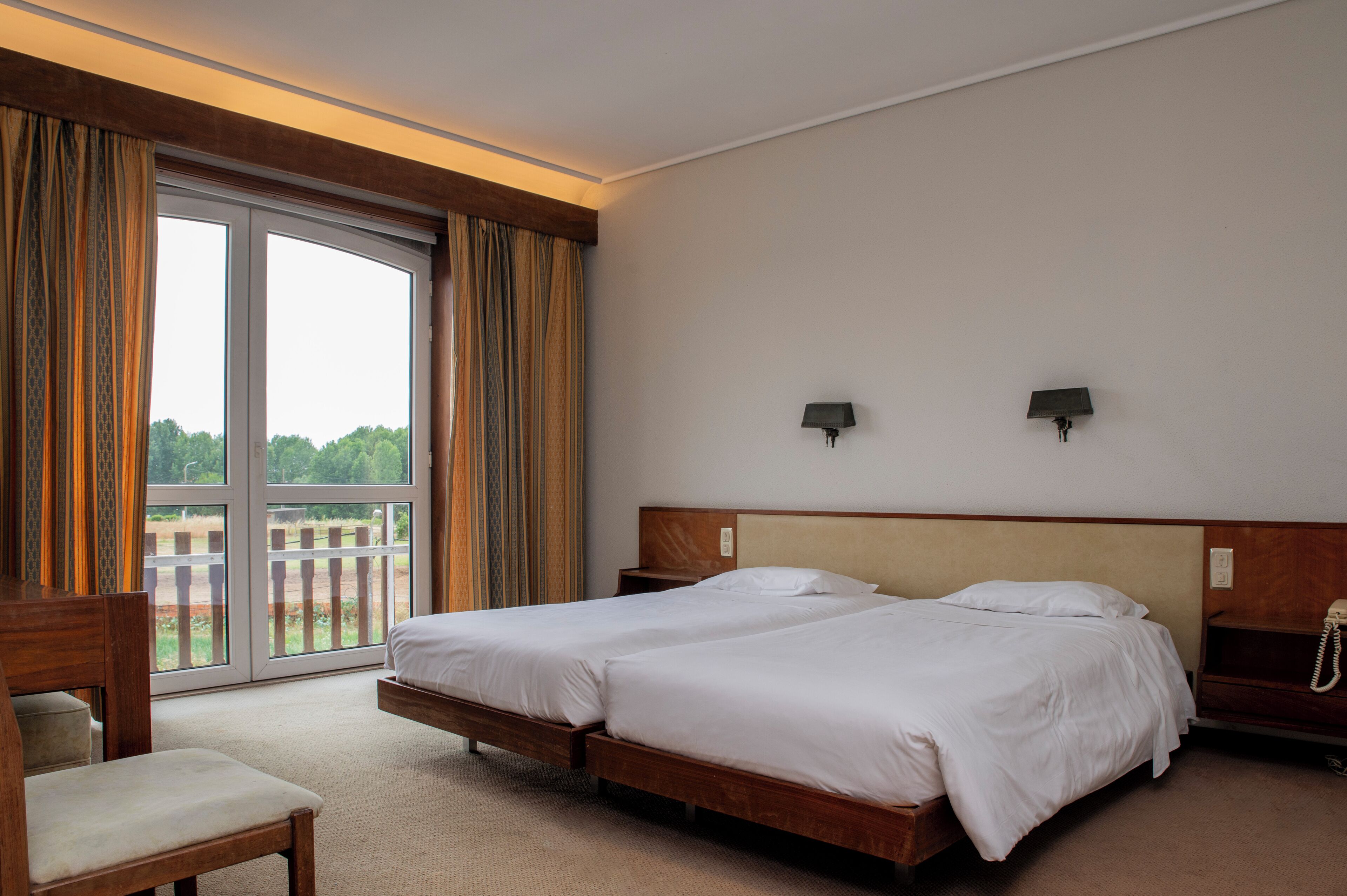 A hotel room with two beds, a wooden headboard, a desk, a chair, a large window with curtains, and a view of greenery outside.