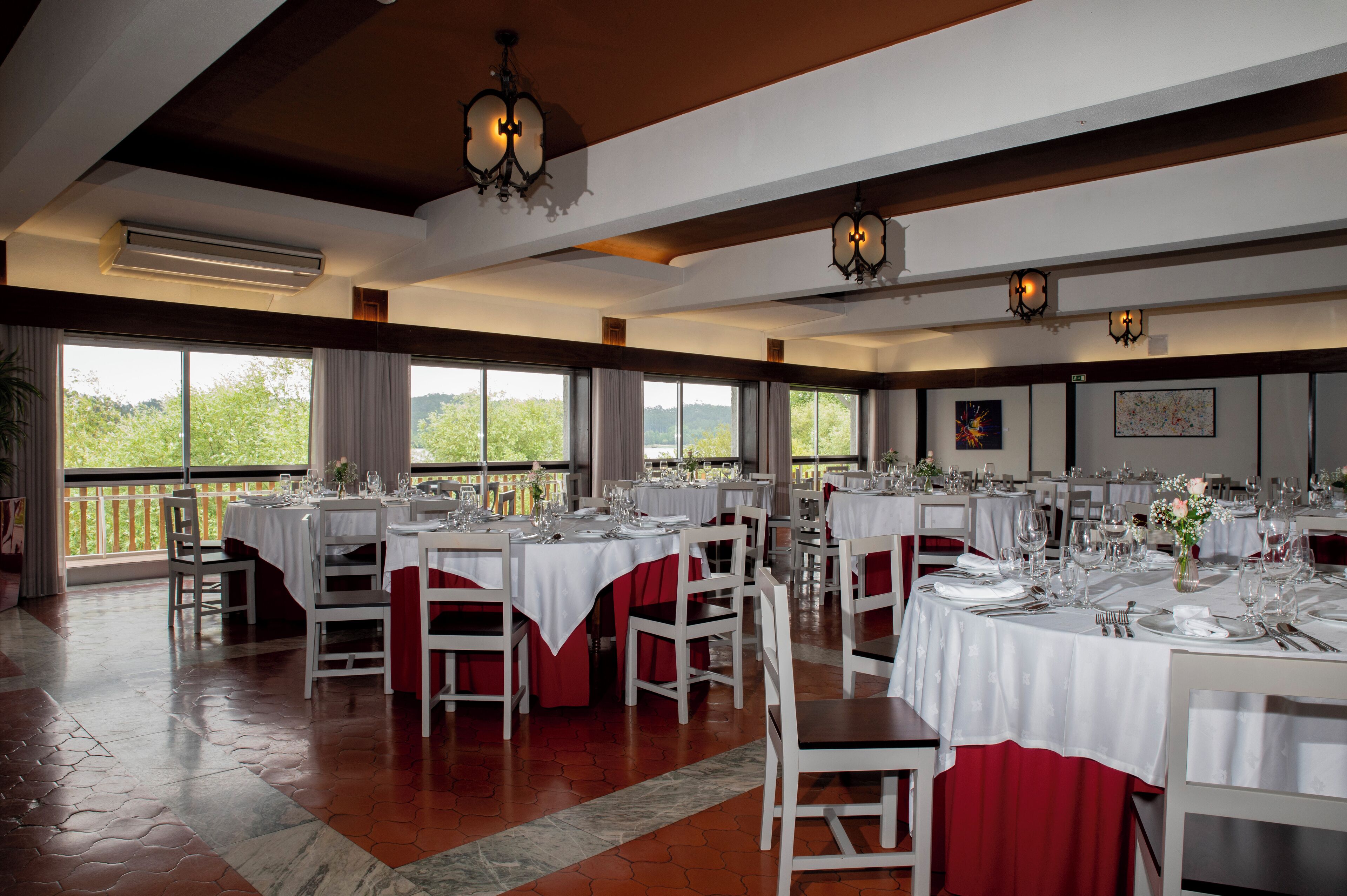 A dining area with multiple tables set for a meal, large windows offering a view of greenery, and a high ceiling with exposed beams.