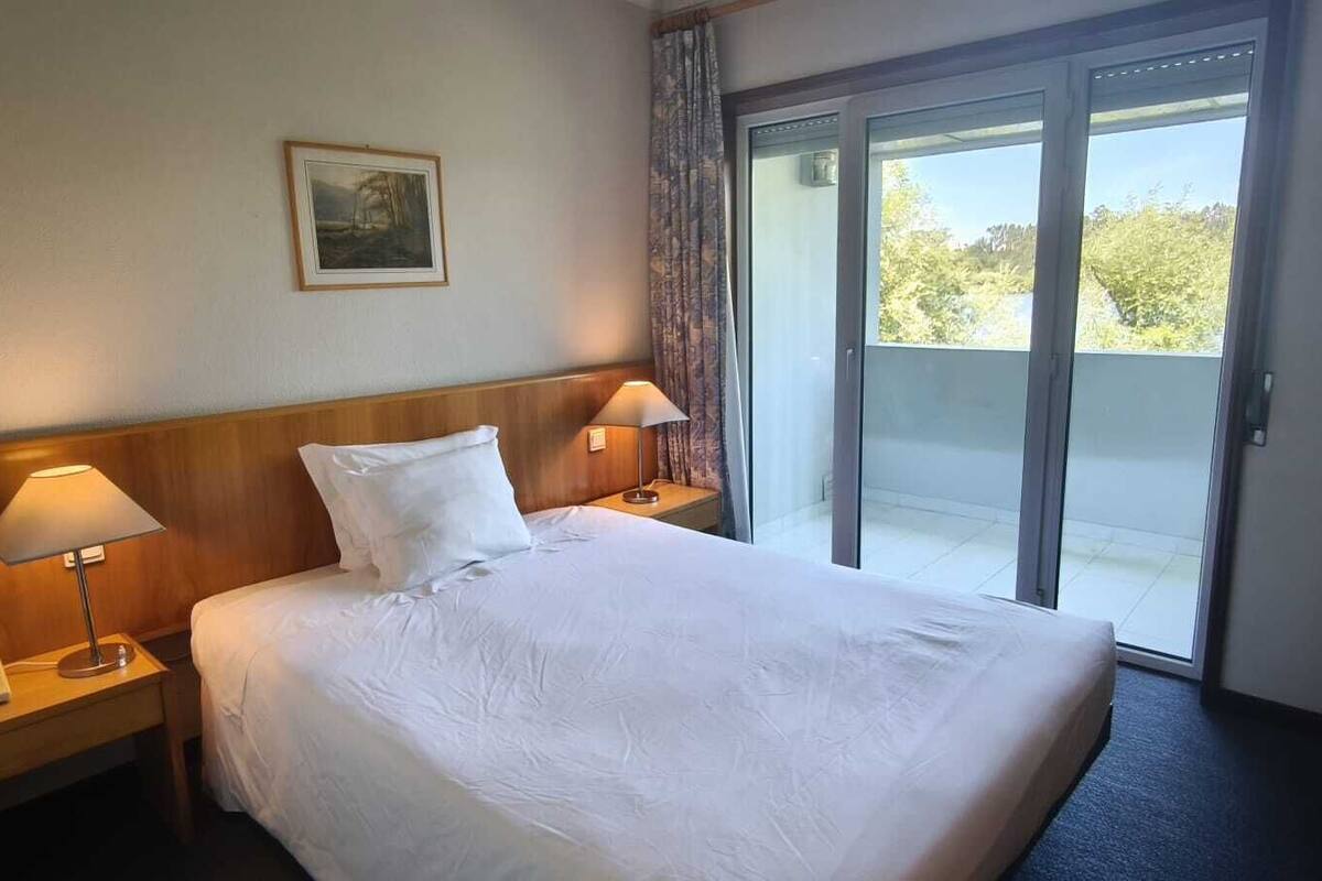 A hotel room with a bed, two bedside lamps, a telephone, a painting on the wall, and a view through a sliding glass door.
