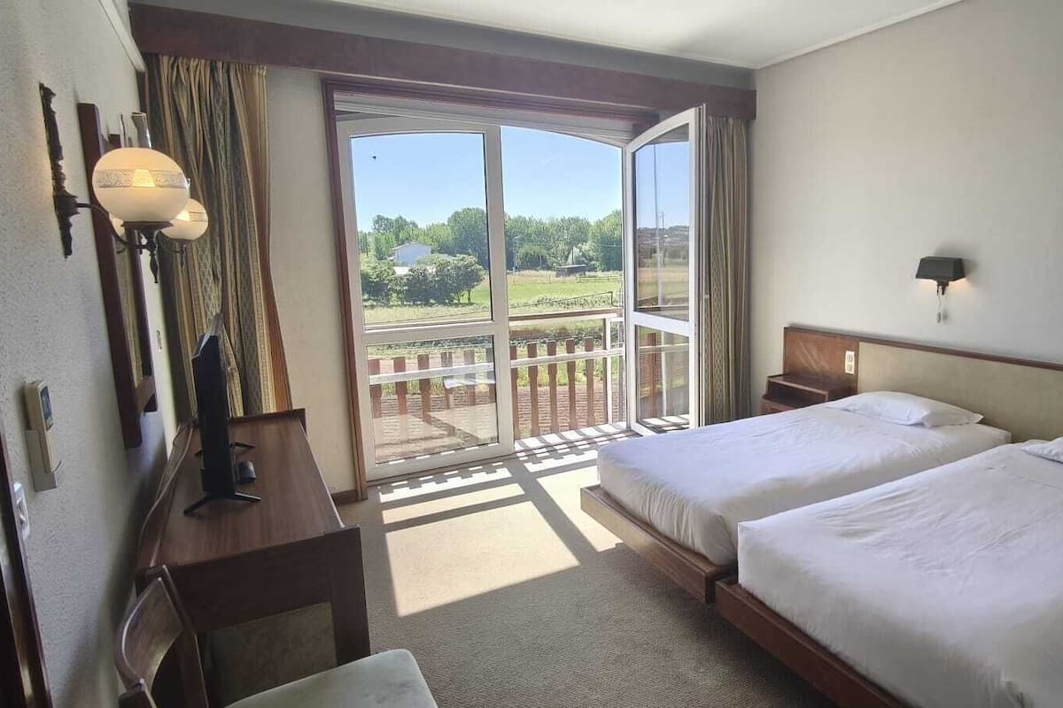 A hotel room with two beds, a desk, a chair, a lamp, a phone, and a balcony with a view of greenery.