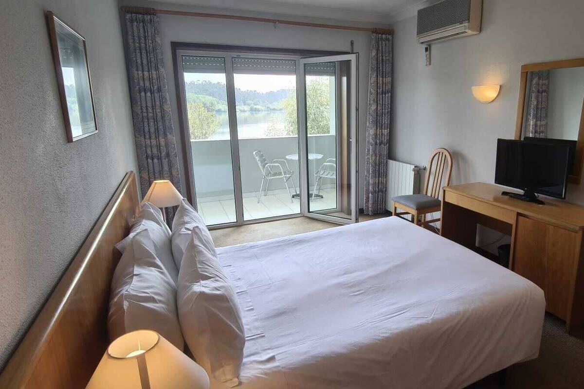 A hotel room with a bed, a desk with a TV, a chair, and a view of the outdoors through a sliding glass door.