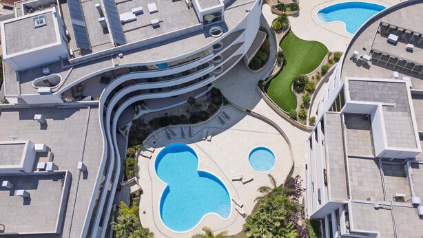 An aerial view of a modern hotel with multiple swimming pools, curved architecture, and landscaped areas.