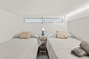 Une petite chambre bien éclairée, avec deux lits simples, une table de chevet avec une lampe et un petit meuble de rangement.