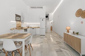 A modern kitchen with white cabinetry, a dining area with a wooden table and chairs, and a hallway with a wooden cabinet and a lamp. - Apartment 'Ilusión' with Private Terrace, Wi-Fi and Air Conditioning (Fuengirola)