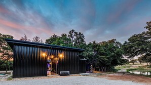 A rustic building with a metal exterior, a gravel path leading to it, and a pond in the background. - Wooded Retreat w/ Pond View near LBJ Grasslands - 4 Cabins/Sleeps 8 (Alvord)