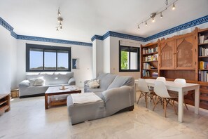 A living room with a sofa, a coffee table, a dining table with chairs, a bookshelf, and a window with a view. - Bahia Golf Suites apartment overlooking the Bay of Cadiz with private pool and terrace (El Puerto de Santa María)